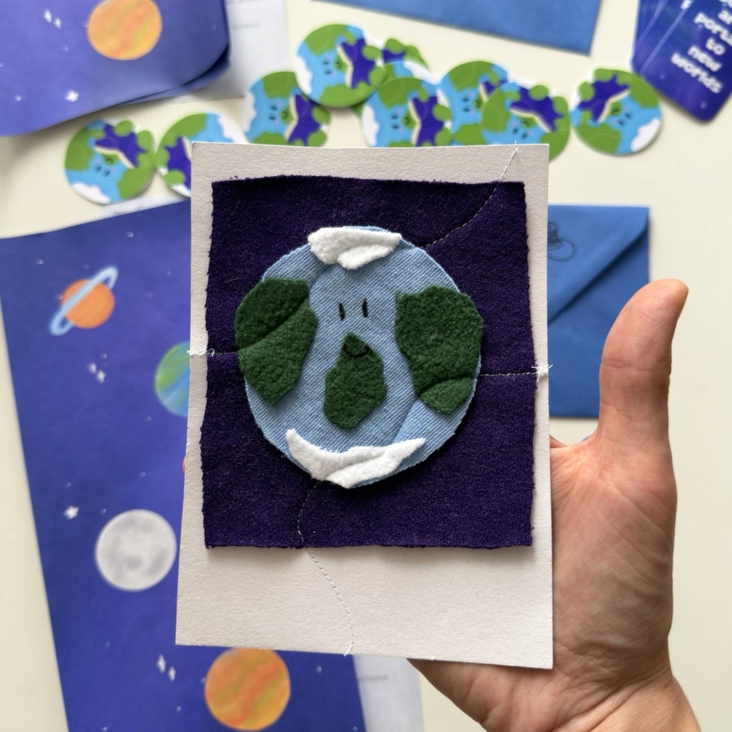 A hand holds a handmade felt planet Earth craft card against a tabletop with matching blue and purple space-themed cards and small planet cutouts.